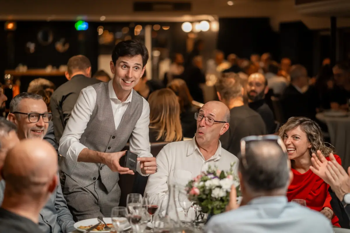 Cyril Regard à table, une boîte de cartes à la main : un invité bouche bée, une invitée éclate de rire